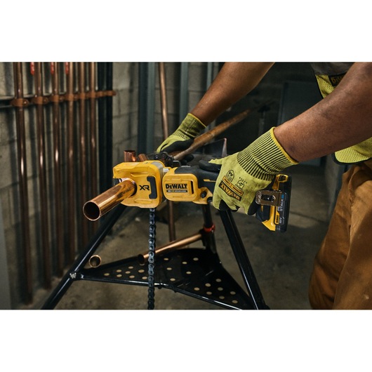 A person wearing green work gloves using a yellow DEWALT cordless power tool to cut a copper pipe, with the tool fixed on a stand in a workshop.