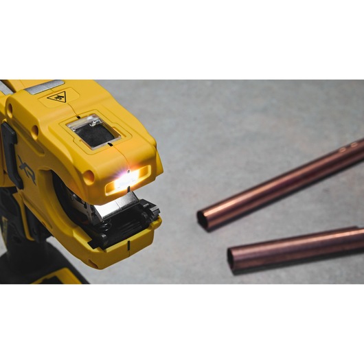 Close-up image of a yellow DEWALT power tool with a built-in light, positioned near copper pipes on a grey surface.