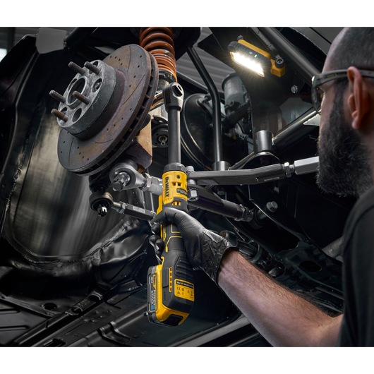 A person wearing gloves uses a DEWALT DCF512EN cordless ratchet tool to perform maintenance on a car's suspension and brake assembly. An LED work light is illuminating the area under the vehicle.
