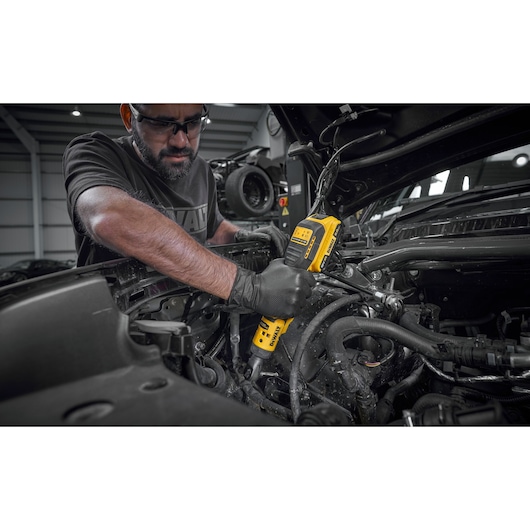A person wearing black gloves is using a DEWALT DCF514N cordless power tool to work on the engine area of a vehicle. The tool is yellow and black, and the scene is set in an automotive workshop.