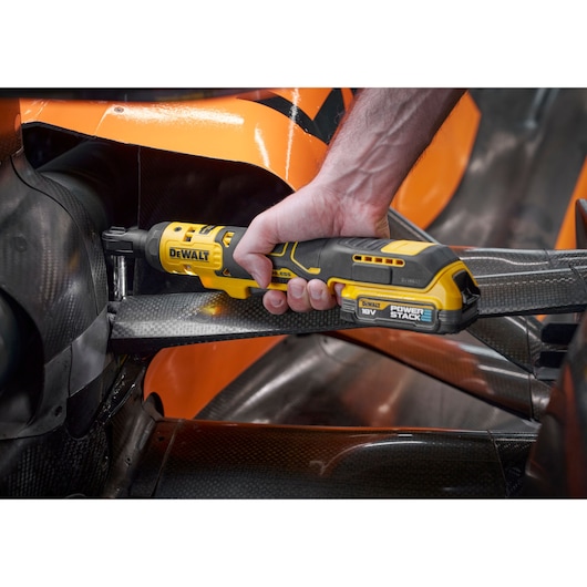 A close-up of a person using a DEWALT DCF514N 18V cordless ratchet wrench with POWERSTACK battery to work on a component of an orange and black machine, possibly a vehicle. The tool is being used in a tight, hard-to-reach space.