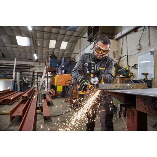 A person wearing protective gloves uses a DEWALT cordless angle grinder to cut a metal bar, producing sparks in an industrial workshop setting.