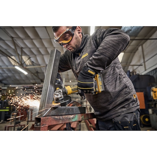 A person wearing DEWALT gloves and work clothes is using a DEWALT DCG404S2T cordless angle grinder to cut a metal beam, creating sparks, in an industrial workshop setting.