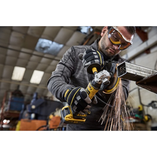A person wearing protective gloves uses a yellow DEWALT cordless angle grinder to cut through a metal pipe in an industrial workshop, with sparks flying from the cutting area.