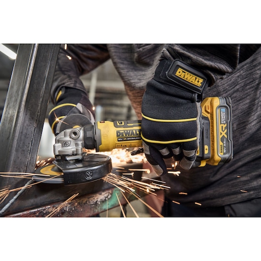 A close-up view of a person wearing gloves using a DEWALT cordless angle grinder on a piece of metal, with visible sparks flying during the grinding process.