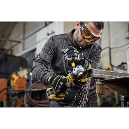 A person wearing protective gloves uses a DEWALT cordless angle grinder to cut a metal pipe, with sparks flying. The scene takes place in an industrial workshop.