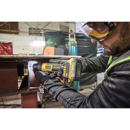 A person wearing gloves uses a DEWALT DCG420N cordless power tool to grind or polish metal inside an industrial workshop.