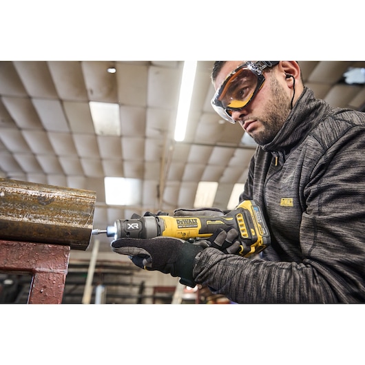 A person wearing a DEWALT jacket and gloves is using a DEWALT cordless rotary tool (model DCG420N) to work on a piece of metal in an industrial workshop.
