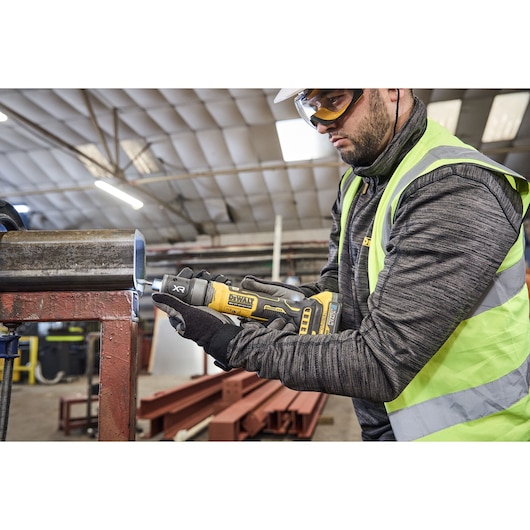 A person wearing safety gear is using a DEWALT DCG420N power tool to work on a metal pipe in an industrial setting.