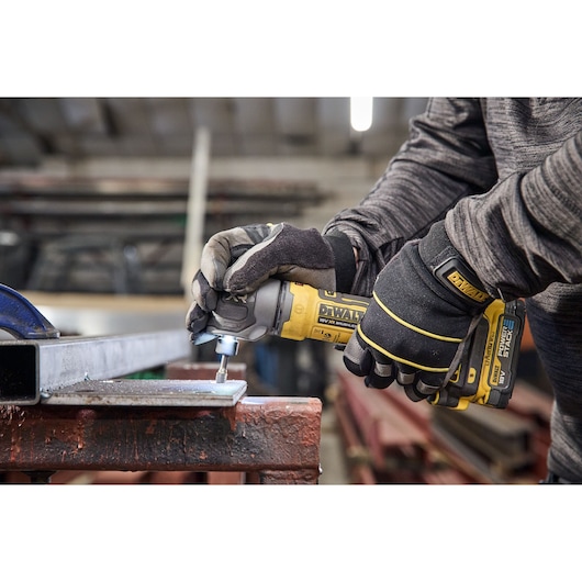 A person wearing DEWALT gloves is using a DEWALT cordless tool to grind or polish a piece of metal in an industrial workshop setting.
