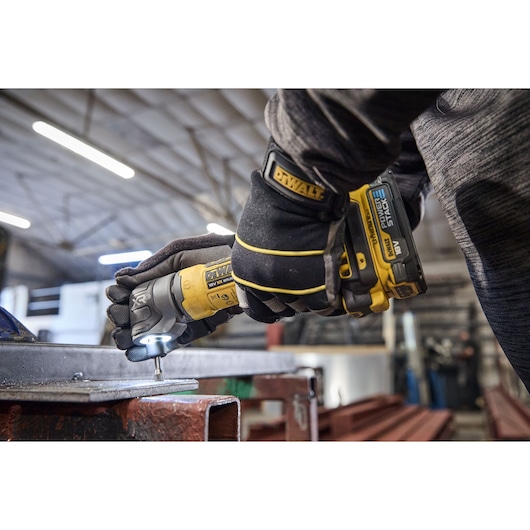 A close-up of a person wearing black and yellow DEWALT branded gloves using a DEWALT cordless power tool to fasten a screw into a metal surface inside an industrial workshop.