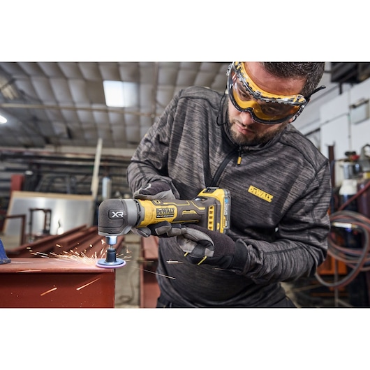 A person wearing safety gloves and a DEWALT jacket is using a DEWALT cordless angle grinder to grind a metal edge, creating sparks in an industrial workshop.