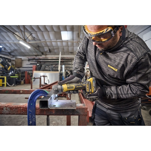 A person uses a DEWALT cordless power tool to drill into a metal object clamped to a workbench in an industrial workshop. The individual's face is obscured for privacy.