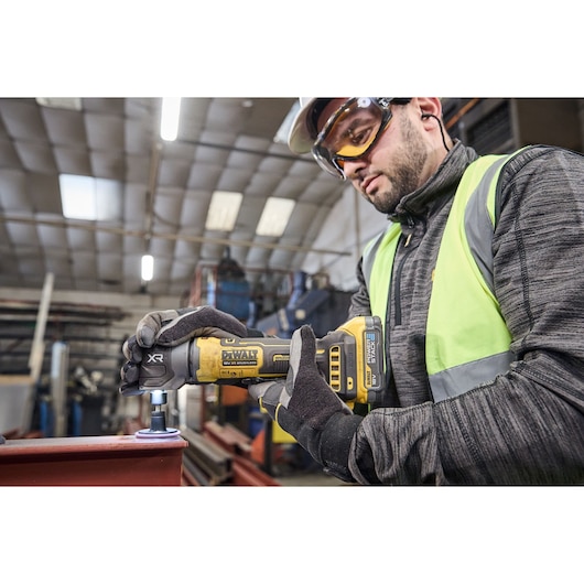 A person wearing gloves and a high-visibility vest is using a DEWALT XR cordless tool on a metal surface inside an industrial workshop. The tool is being operated close to the edge of a red metal beam.
