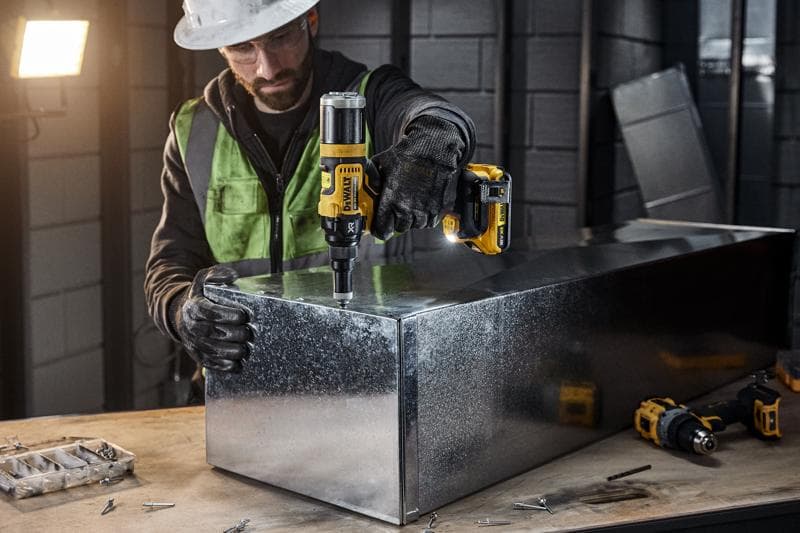 Una persona con attrezzatura di sicurezza utilizza un utensile elettrico senza fili DEWALT DCF403NT per lavorare su una scatola di metallo in un laboratorio industriale. Vari viti e attrezzi sono visibili sul banco da lavoro.