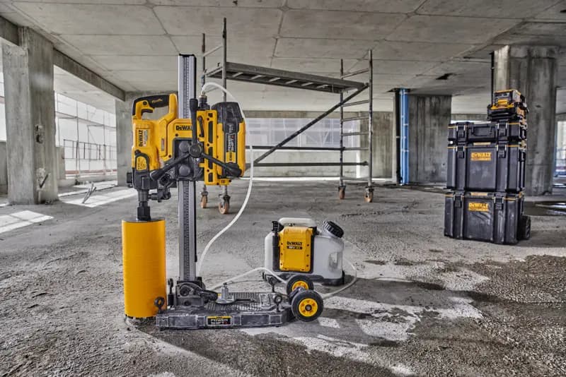 Una macchina per carotaggio DEWALT con accessorio di trapano cilindrico giallo e serbatoio dell'acqua collegato è posizionata sul pavimento di un cantiere in cemento. Sullo sfondo sono visibili un'impalcatura e scatole impilabili DEWALT.