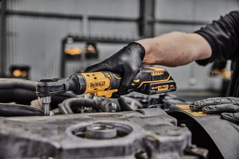 Una persona con guanti neri utilizza uno strumento a cricchetto senza fili DEWALT su un componente del motore in un'officina.