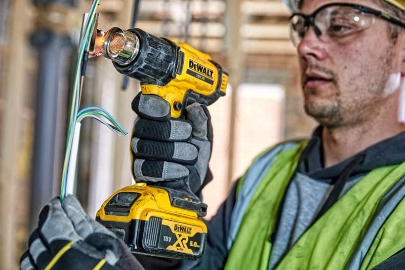 Una persona con guanti da lavoro e un giubbotto di sicurezza utilizza una pistola termica a batteria DEWALT DCE530N per applicare calore ai fili elettrici in un cantiere. Il volto della persona è sfocato per motivi di privacy.