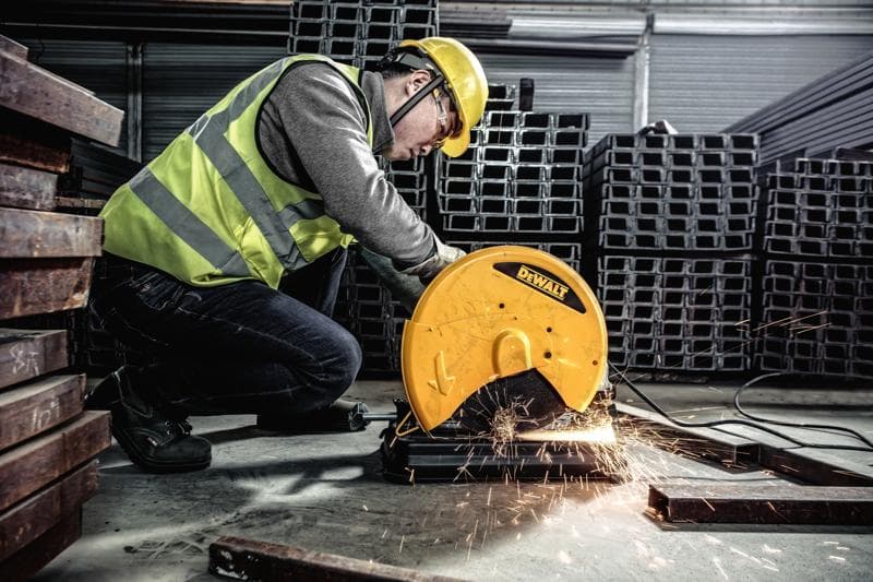 Una persona con indumenti di sicurezza utilizza una sega da taglio per metallo DEWALT, modello D28730, per tagliare barre di metallo in un ambiente industriale. Scintille volano dalla zona di taglio.