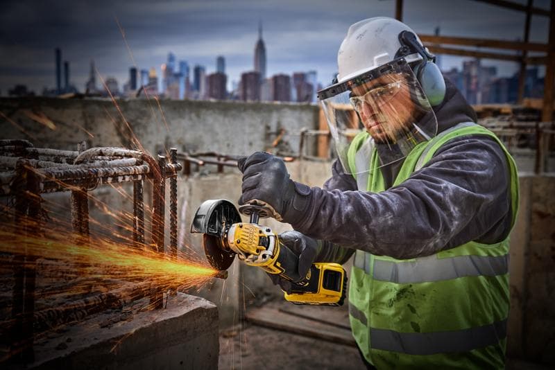Operaio usa smerigliatrice DEWALT DCG405P2 su cantiere, indossa protezioni, città sullo sfondo.