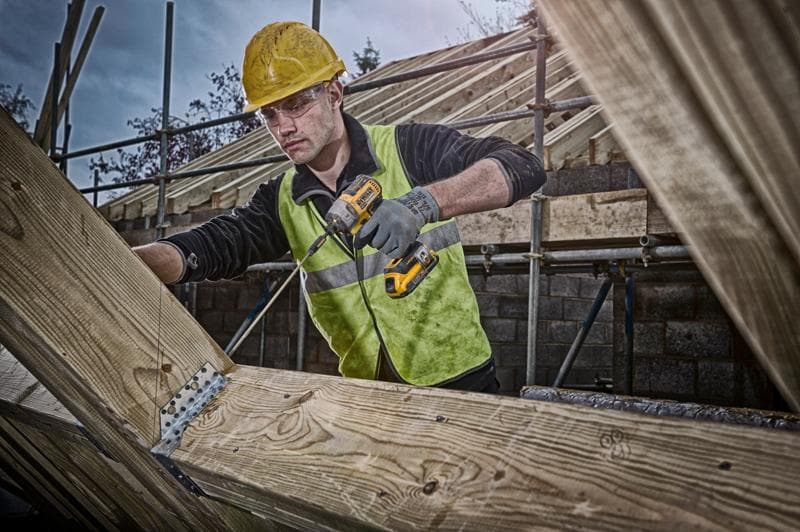 Un operaio edile con casco di sicurezza giallo e giubbotto riflettente utilizza un trapano cordless DEWALT per fissare staffe metalliche su travi di legno in un cantiere. Il suo volto è sfocato.