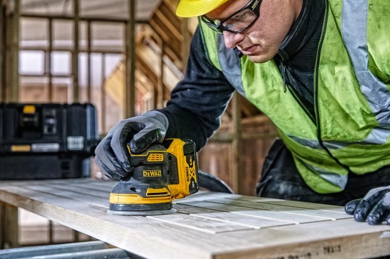 DEWALT DCW210, DCW210D1 levigatrice su legno, scena di cantiere, persona con guanti e giubbotto di sicurezza.