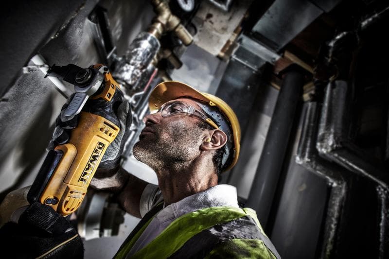 Operaio taglia un tubo a muro con DEWALT DWE315, indossa casco e gilet in ambiente industriale.