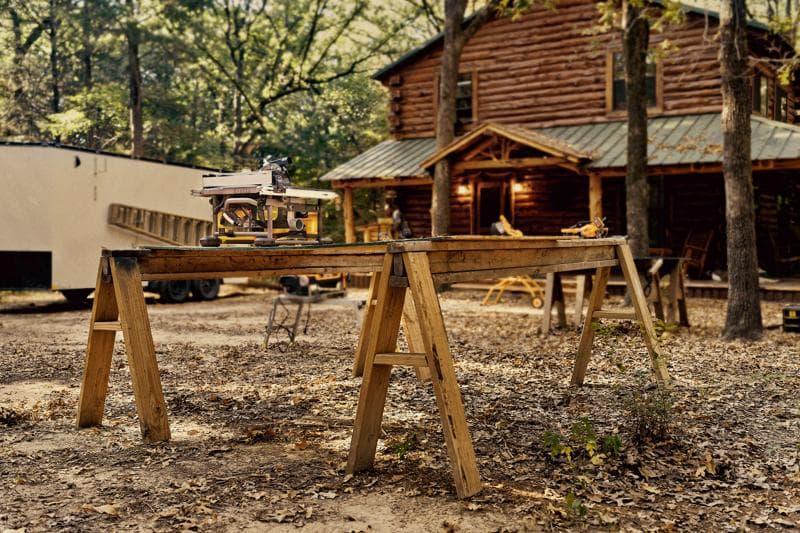 Una sega da banco DEWALT posizionata su un banco da lavoro in legno composto da due cavalletti all'aperto, con una baita in legno e alberi sullo sfondo.