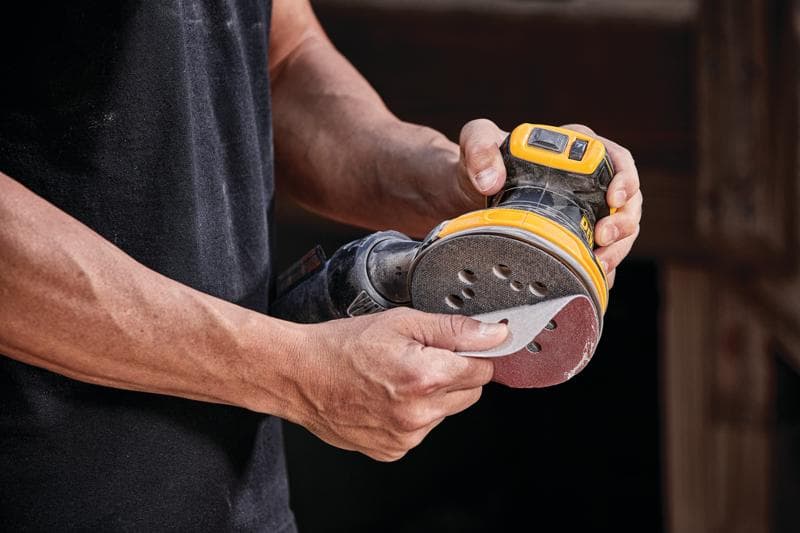 Una persona tiene in mano una levigatrice orbitale casuale a batteria DEWALT, modello DCW210B, fissando un disco abrasivo sull’attrezzo.