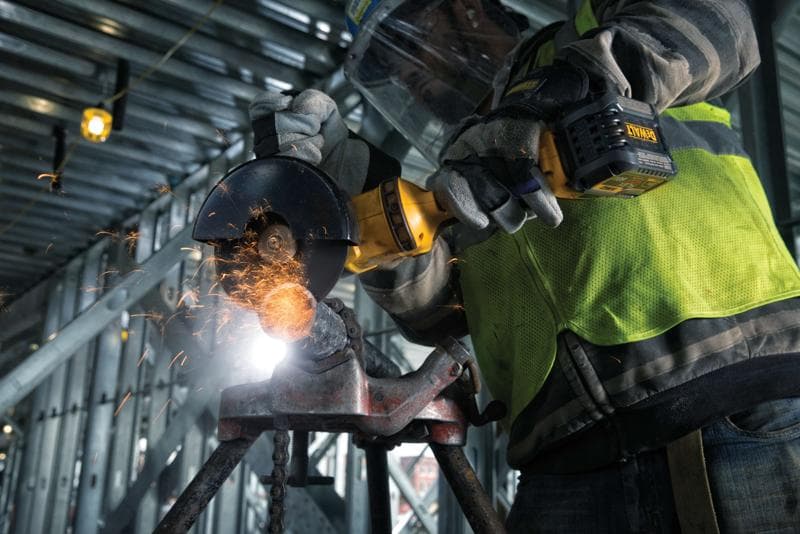 Primo piano di una persona con guanti protettivi e giubbotto di sicurezza che utilizza una smerigliatrice angolare DEWALT senza fili per tagliare metallo, con scintille che volano, all’interno di un cantiere industriale.