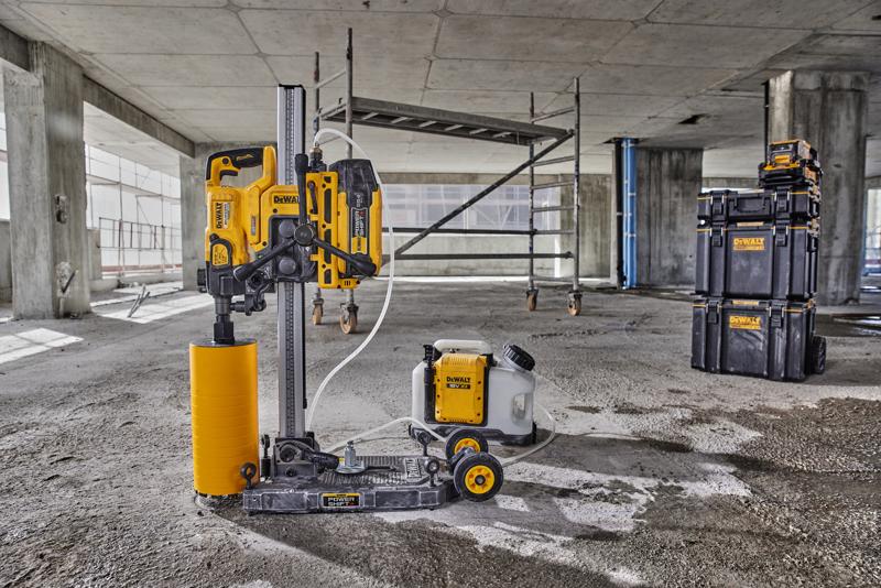 Una macchina per carotaggio DEWALT con accessorio di trapano cilindrico giallo e serbatoio dell'acqua collegato è posizionata sul pavimento di un cantiere in cemento. Sullo sfondo sono visibili un'impalcatura e scatole impilabili DEWALT.