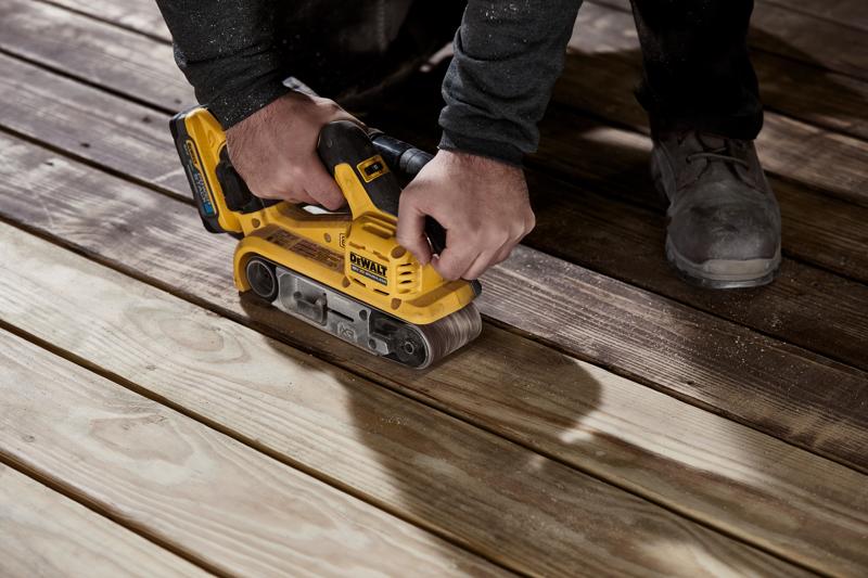 Una persona utilizza una levigatrice a nastro senza fili gialla DEWALT su tavole di legno per levigare la superficie.