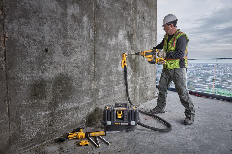 Un operaio edile con gilet di sicurezza e casco sta utilizzando un trapano DEWALT senza fili con accessorio per l'aspirazione della polvere per forare una parete di cemento. L'operaio è su un grande edificio con panorama urbano sullo sfondo. Vari attrezzi DEWALT e una valigetta degli attrezzi sono posati a terra nelle vicinanze.