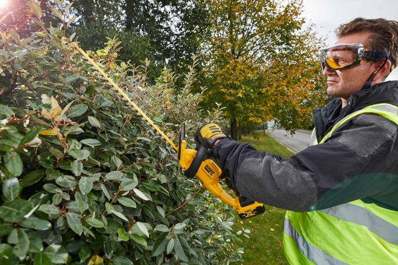 Persona usa DEWALT DCM563PB tagliasiepi all'aperto, tagliando siepi vicino a una strada e a alberi autunnali.