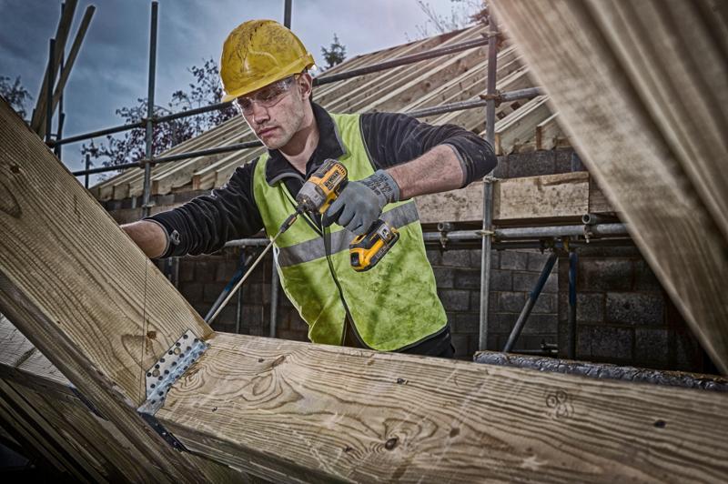 Un operaio edile con casco di sicurezza giallo e giubbotto riflettente utilizza un trapano cordless DEWALT per fissare staffe metalliche su travi di legno in un cantiere. Il suo volto è sfocato.
