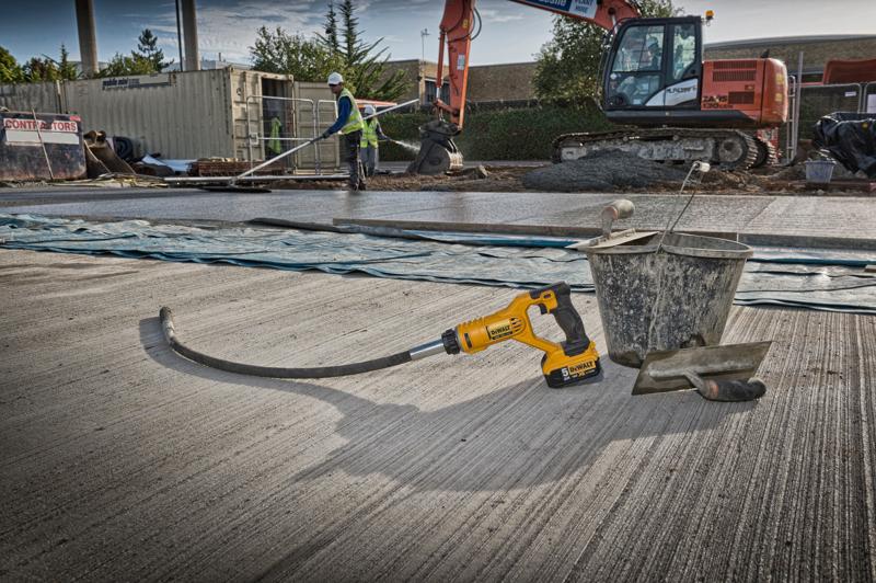 Un vibratore per calcestruzzo DEWALT DCE531N è posizionato su un cantiere, accanto a un secchio di metallo e una spatola su una superficie di cemento appena gettata. Sullo sfondo ci sono operai edili, un grande escavatore e altri materiali da costruzione.