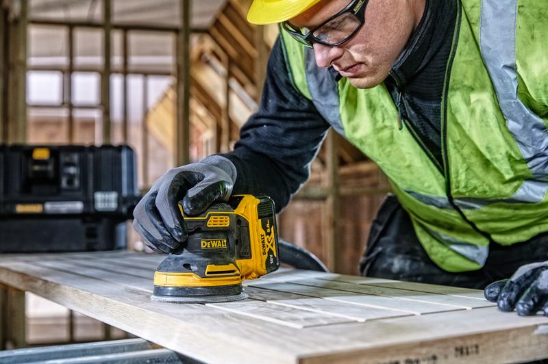DEWALT DCW210, DCW210D1 levigatrice su legno, scena di cantiere, persona con guanti e giubbotto di sicurezza.