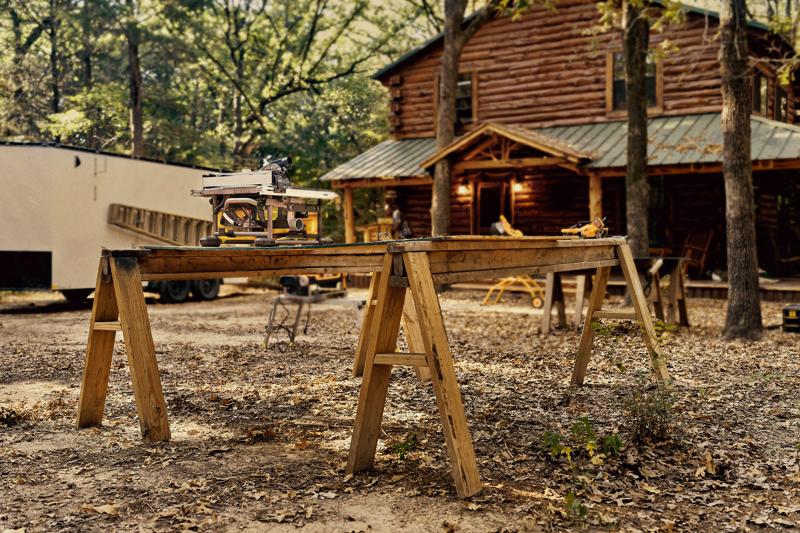 Una sega da banco DEWALT posizionata su un banco da lavoro in legno composto da due cavalletti all'aperto, con una baita in legno e alberi sullo sfondo.