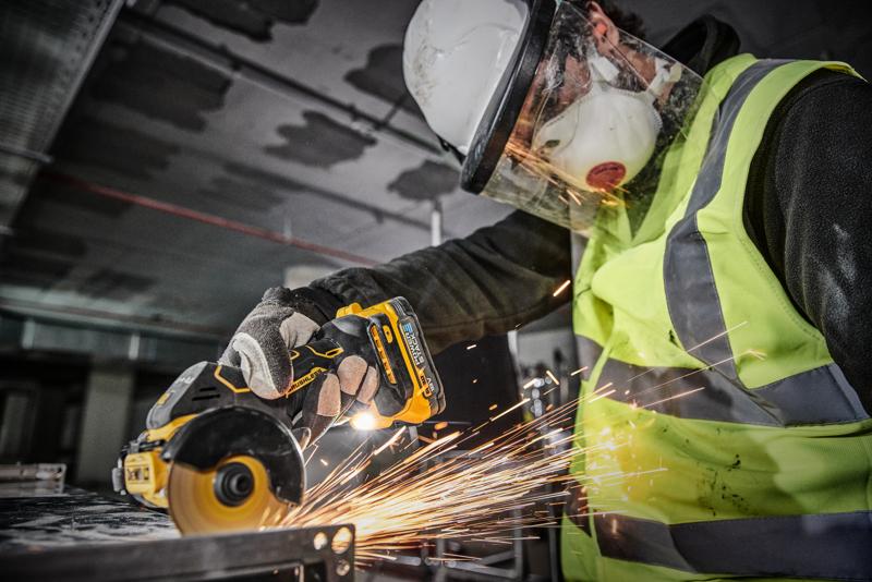 Una persona indossa dispositivi di sicurezza, tra cui casco, visiera, mascherina, guanti e gilet ad alta visibilità, e utilizza un utensile elettrico a batteria DEWALT DCS438N per tagliare metallo, generando scintille.